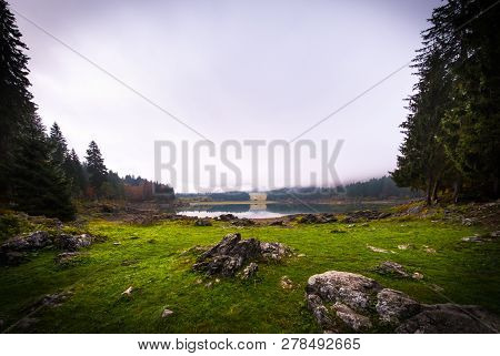 View Over Lake Laghi Di Fusine On A Foggy Morning With Some Rocks At The Beach And Mountain Range Ma