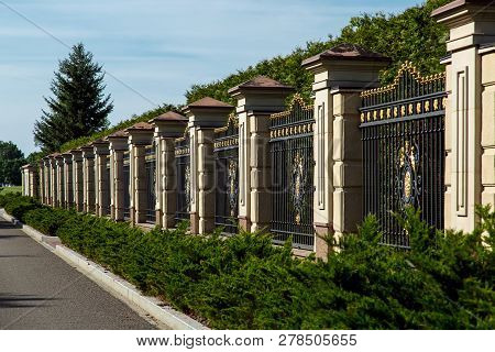 A Fence With Columns And A Lattice With A Forged Pattern, A Fence Near The Asphalt Road And Bushes O