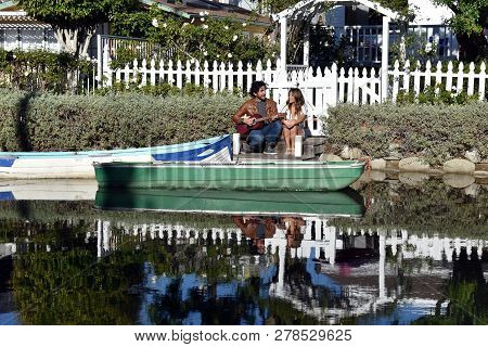 Handsome Young Man Playing Guitar For A Pretty Girl By A Beautiful Canal With Boats