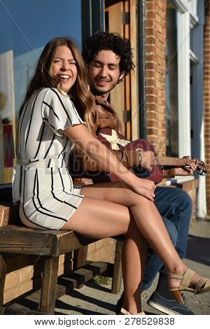 A Young Man Playing The Guitar For A Pretty Girl Outdoors On A Bench