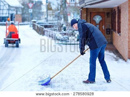 Man With Snow Shovel Cleans Sidewalks In Winter During Snowfall. Winter Time In Europe. Young Man In