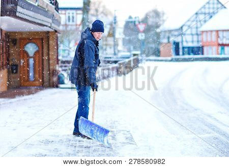 Man With Snow Shovel Cleans Sidewalks In Winter During Snowfall. Winter Time In Europe. Young Man In