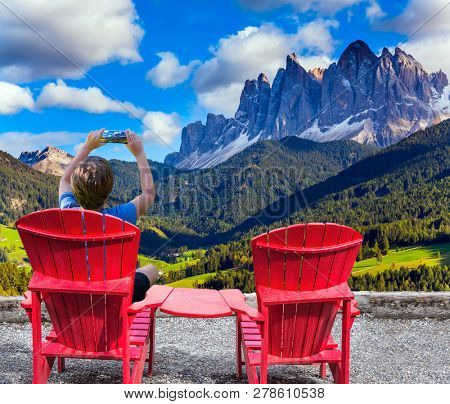 Boy sits in a red lounge chair and photographs a beautiful landscape with a mobile phone. Wonderful day in the Dolomites, South Tyrol. The concept of ecological and photo tourism