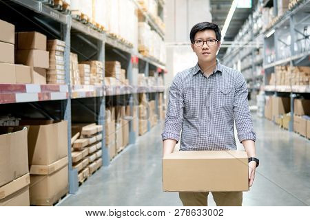 Young Asian Man Carrying Cardboard Box Between Row Of Shelves In Warehouse. Shopping Warehousing Or 