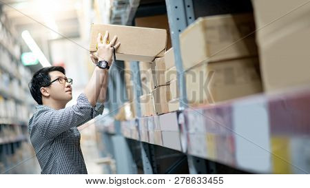 Young Asian Man Taking Cardboard Box From Shelves In Warehouse. Shopping Warehousing Or Working Pick