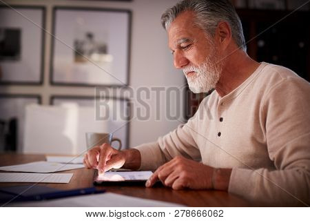 Senior Hispanic man sitting at a table using a tablet computer  in the evening, close up, side view