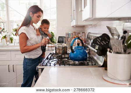 Multitasking mum holding her young baby while she makes food at the hob in her kitchen, side view