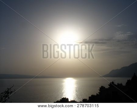 Impressive silver sunset at the Adriatic coast with the sun reflected in the sea, mountains at the distance and a cloudlet in a clear sky.