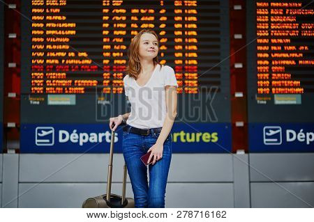 Young Woman In International Airport With Luggage And Passport Near Flight Information Display