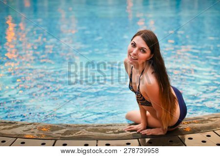 Sexy Woman Sitting On The Edge Of Swimming Pool, Wearing Bikini While On Vacations In A Sunny Tropic