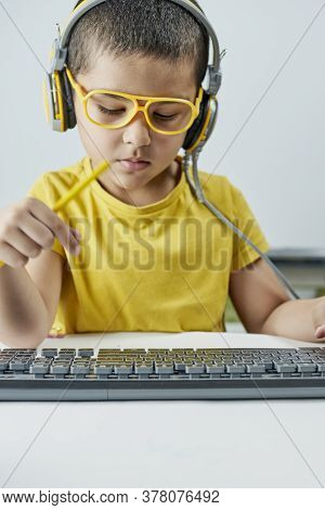 An Adorable Kid In A Yellow T-shirt With A Headphone Listening Online Course. Boy Typing On Pc Keybo