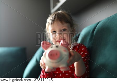 Girl In Red Dress With Polka Dots Sit On Couch And Smile. Child Hold Pink Piggy Bank In Her Hands An