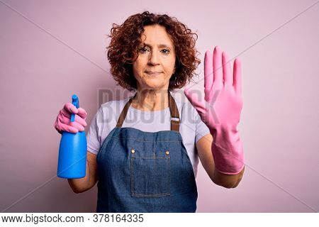 Middle age curly hair woman cleaning doing housework wearing apron and gloves using spayer with open hand doing stop sign with serious and confident expression, defense gesture