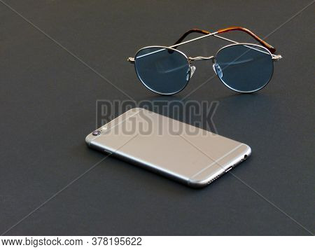 Glasses In Gray Frames With Blue Lenses Next To A Gray Phone On A Black Background