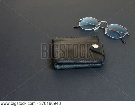 Glasses In Gray Frames With Blue Lenses Next To A Black Leather Wallet On A Black Background