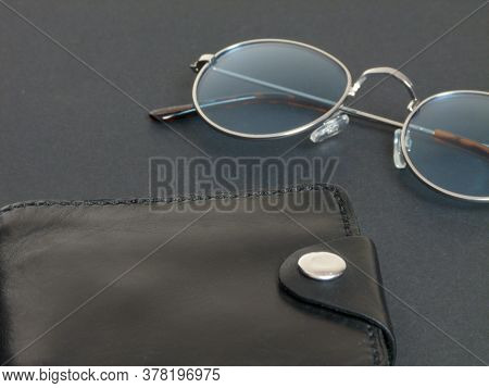 Glasses In Gray Frames With Blue Lenses Next To A Black Leather Wallet On A Black Background