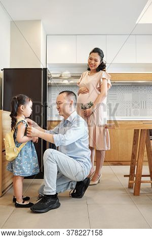 Smiling Pregnant Vietnamese Woman Looking At Her Husband Helping Little Daughter To Wear Backback Wh