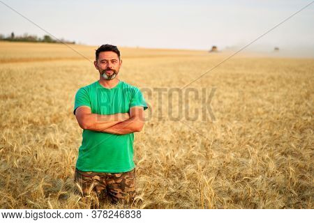 Happy Farmer Proudly Standing In Wheat Field With Arms Crossed On Chest. Agronomist Wearing Corporat