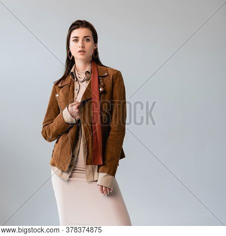 Attractive, Fashionable Girl Touching Suede Jacket While Looking At Camera Isolated On Grey