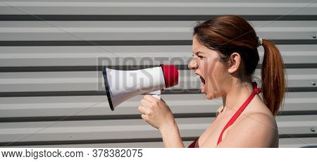 Red-haired Caucasian Woman In A Red Bikini Shouting Through A Megaphone Against A Background Of A Gr
