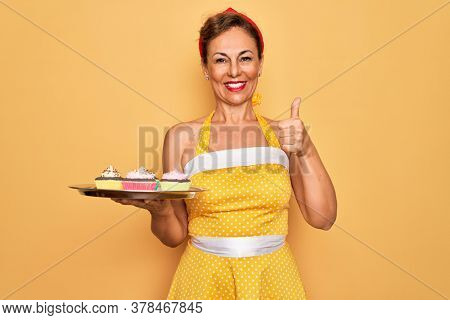 Middle age senior pin up woman wearing 50s style retro dress holding tray with cupcakes happy with big smile doing ok sign, thumb up with fingers, excellent sign