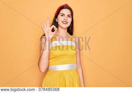 Young beautiful pin up woman wearing 50s fashion vintage dress over yellow background smiling positive doing ok sign with hand and fingers. Successful expression.