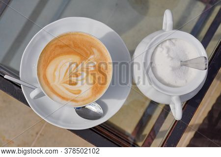 Cup Of Coffee With Milk And Flower As Decoration, Sugar With Small Spoon In White Tableware On Glass