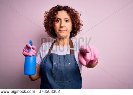 Middle age curly hair woman cleaning doing housework wearing apron and gloves using spayer pointing with finger to the camera and to you, hand sign, positive and confident gesture from the front