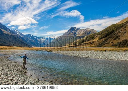 A Fly Fisherman Casting For Trout, Surrounded By Snow Capped Mountains, On The Ahuriri River, South 