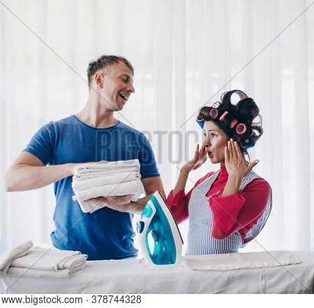 Young Couple At Home Doing Household Chores And Ironing. Wife Ironing Clothes For Her Husband At Wor