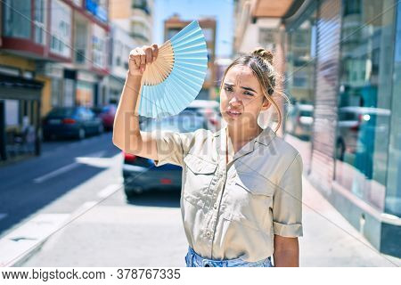 Young beautiful blonde caucasian woman smiling happy outdoors on a sunny day using handfan for hot weather