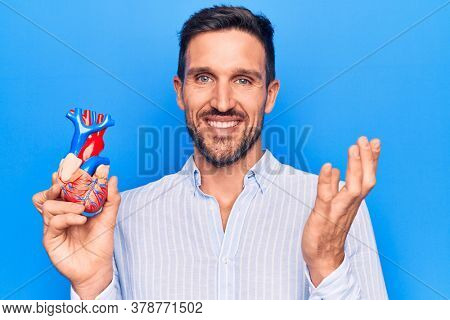 Young handsome man holding heart organ with veins and arteries over blule background celebrating achievement with happy smile and winner expression with raised hand