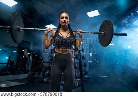 From Below View Of Female Bodybuilder With Long Braids Holding Barbell On Shoulders And Looking At C