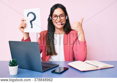 Young caucasian woman sitting at the desk wearing call center agent headset holding question mark smiling happy and positive, thumb up doing excellent and approval sign 