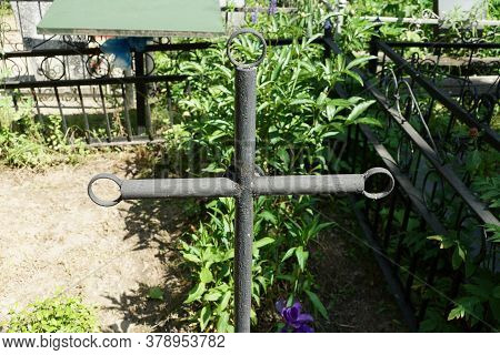 One Black Iron Cross On A Grave In A Cemetery Overgrown With Green Vegetatio