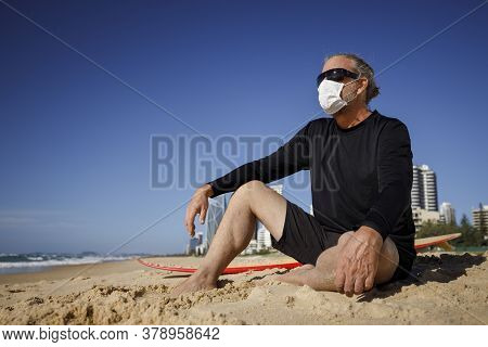 Man With Medical Mask Sitting On Beach During Coronavirus Covid 19 Pandemic, Social Distance Concept