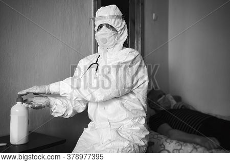 Black And White Image Of Doctor In Ppe Suit Visiting Patient At Home, Decontaminating Hands With Alc