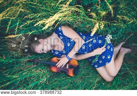 Young Slim Fair-skinned Girl Blonde Beautiful Lies In Tall Grass. A Woman Hugs Violin.