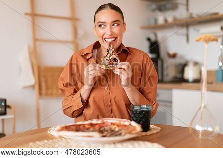 The Joy Of Pizza. Excited Lady Enjoying Eating Tasty Pizza Holding And Biting Slice, Sitting In Kitc