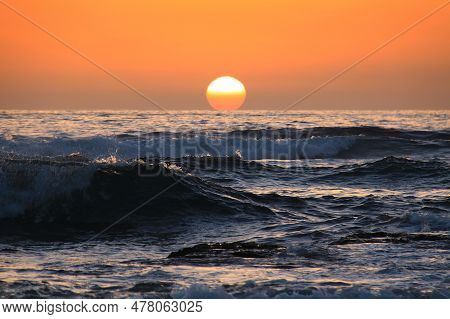 The Sun In An Orange Sky Setting Behind The Sea Waves.