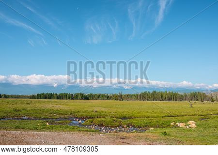 Scenic Landscape With Creek Against Forest And Large Snowy Mountain Range In Low Clouds In Bright Su
