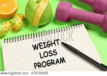 Weight Loss Concept. Notebook, Dumbbells And Apple With Measuring Tape On Green Background, Closeup