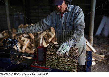 Caucasian Senior Man Splits Wood With A Hydraulic Log Splitter. He Wears A Denim Work Shirt, Work Gl