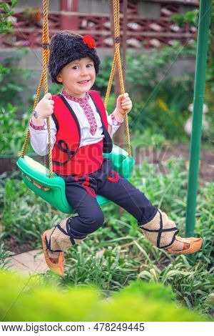 Bulgarian Boy In Ethnic Folklore Costume With Traditional Embroidery On A Swing In A Garden At His  