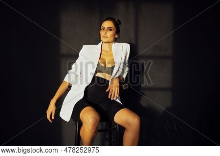Young Pregnant Brunette In White Jacket And Black Shorts Is Sitting On Bar Stool Smiling And Posing.