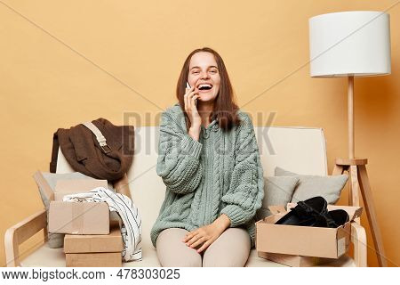 Attractive Laughing Woman Sitting On Sofa Among Boxes With Clothing Against Beige Wall Talking On Sm