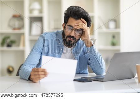Upset Young Indian Man Sitting At Desk With Laptop And Checking Documents