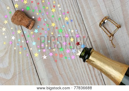 Overhead of a bottle of champagne with the cork popping on a rustic wood table. The spray from the bottle is stars of various sizes. With copy space it is perfect for New Years projects.