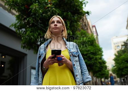 Gorgeous young woman holding mobile phone while standing in the city with beautiful trees