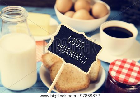 a chalkboard with the text good morning and a blue wooden table with a bottle of milk, a cup of coffee, some toasts in a plate, a jar of jam and a bar of butter